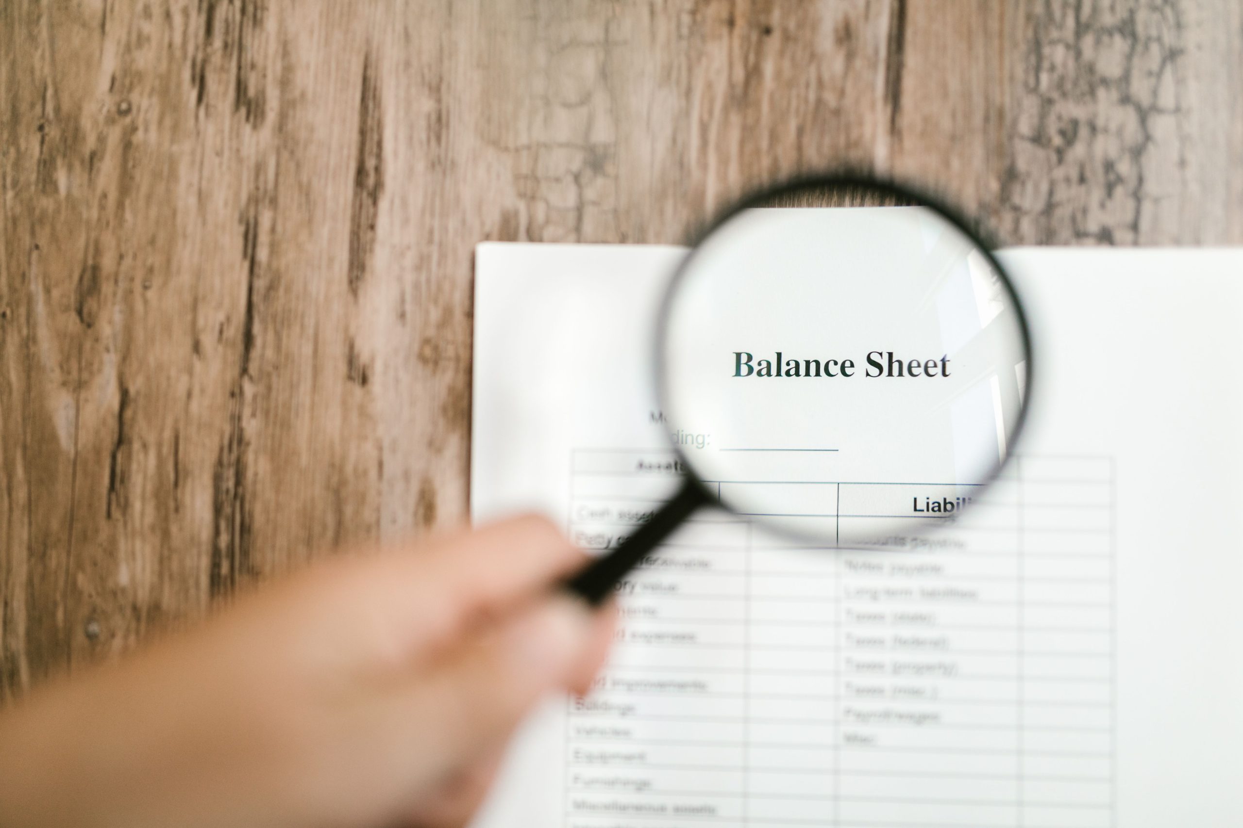 Close-up of a balance sheet document on a wooden surface with a magnifying glass held by a hand