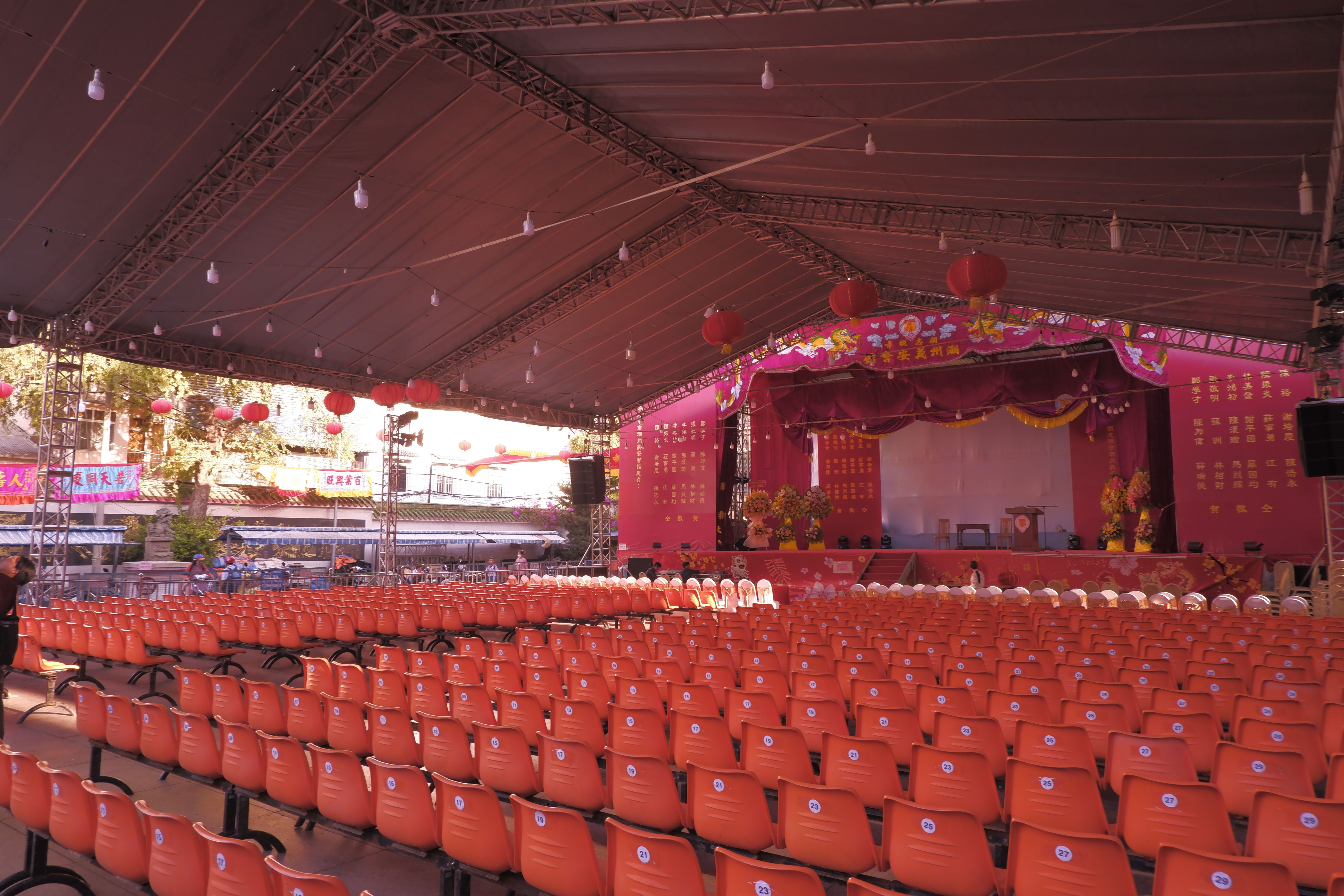 Outdoor performance venue with rows of orange seats facing a decorated stage surrounded by greenery.
