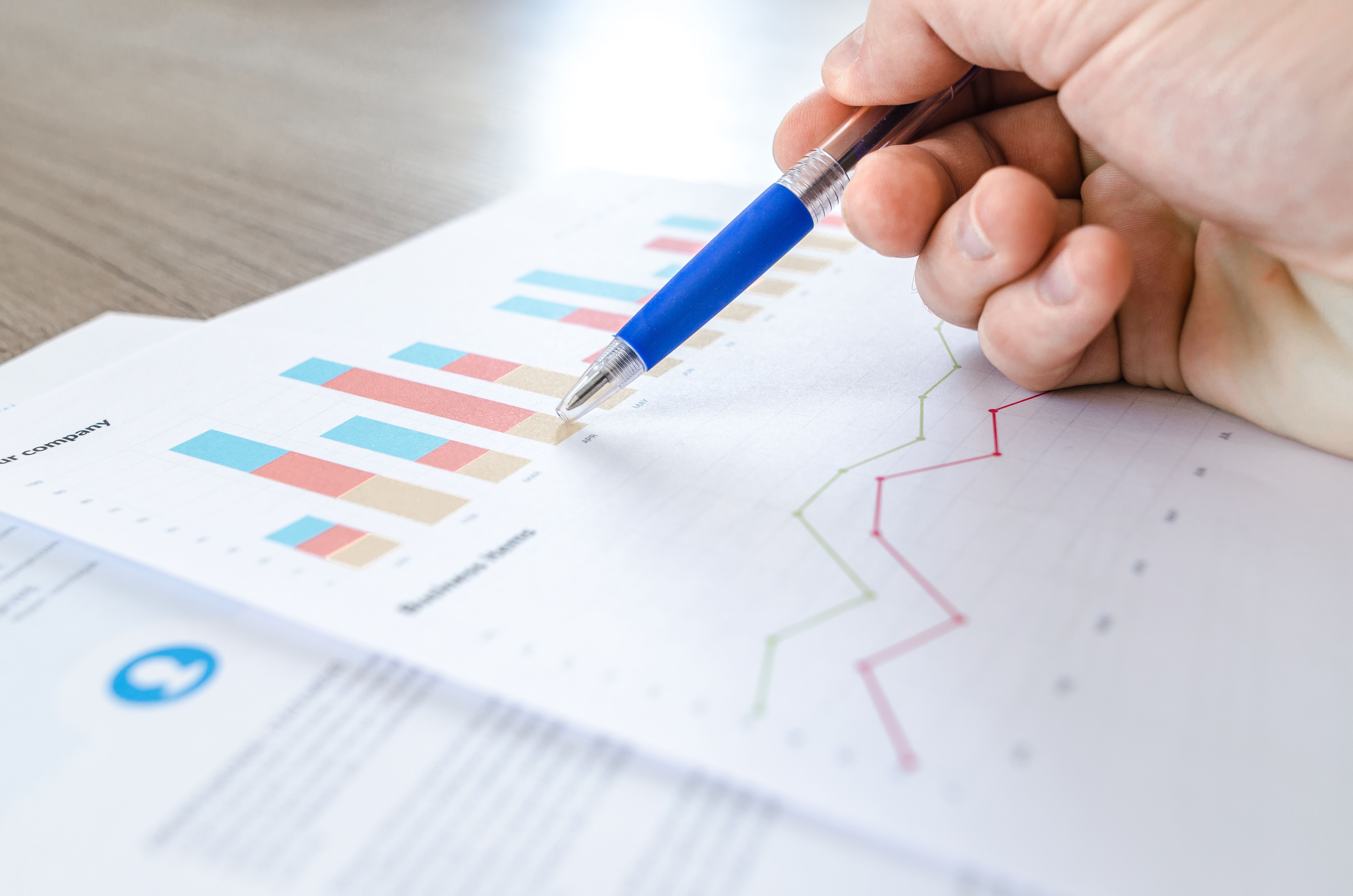 A close-up of a hand with a pen analyzing data on colorful bar and line charts on paper